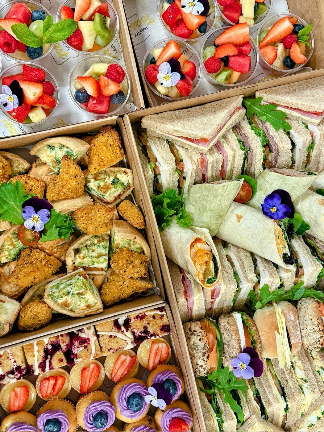 Catering boxes of sandwiches, wraps, savouries, fruit pots and cakes, arranged and garnished with edible flowers